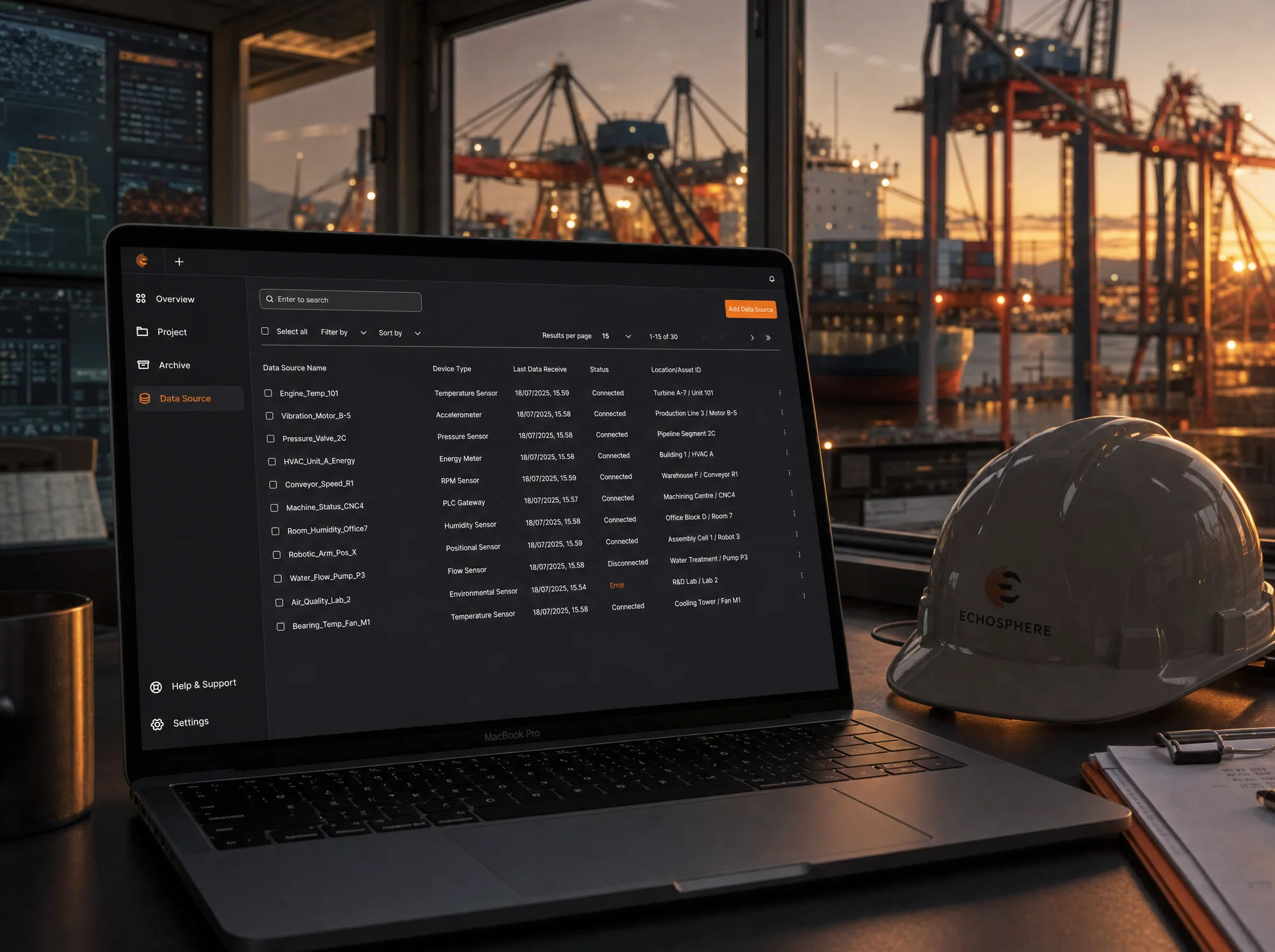 A MacBook Pro in an industrial control room showing the SpatialWorks data source dashboard, shipyard cranes visible through the window behind it